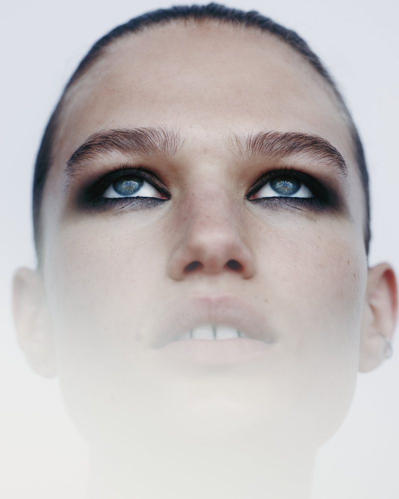 Close-up portrait of a woman with blue eyes and dark eyeshadow, looking up against a white background with soft, diffused lighting.