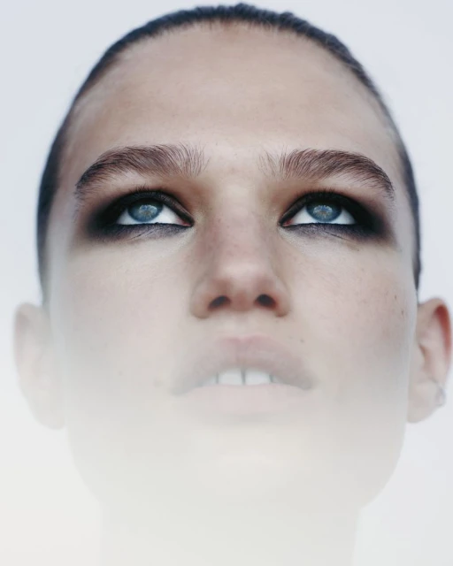 Close-up portrait of a woman with blue eyes and dark eyeshadow, looking up against a white background with soft, diffused lighting.