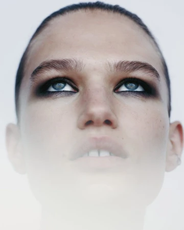 Close-up portrait of a woman with blue eyes and dark eyeshadow, looking up against a white background with soft, diffused lighting.