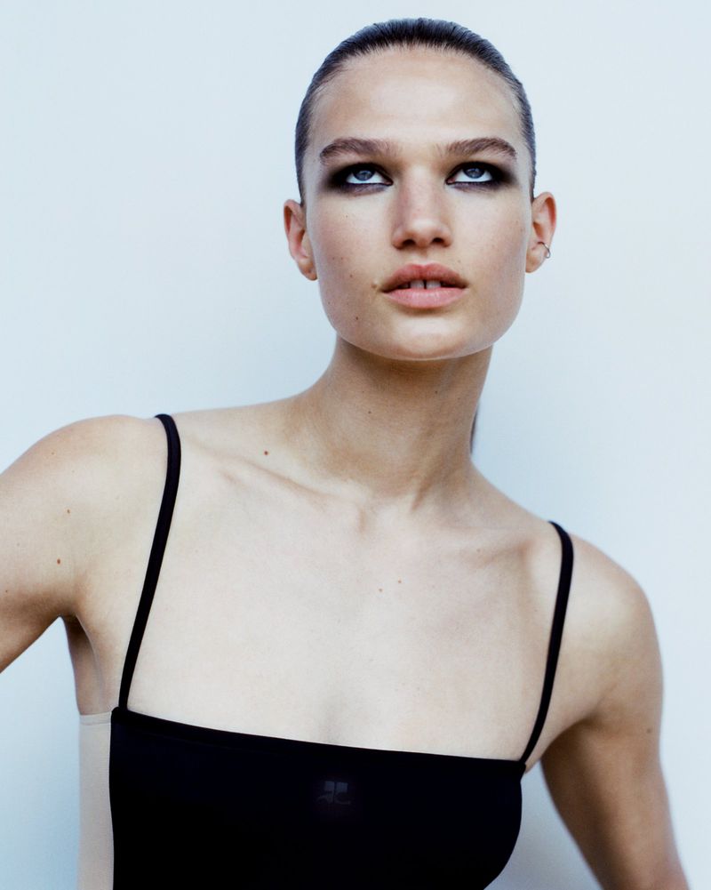 Portrait of a young adult female fashion model in a simple black top, fair skin, and dark eyeshadow against a plain white background.