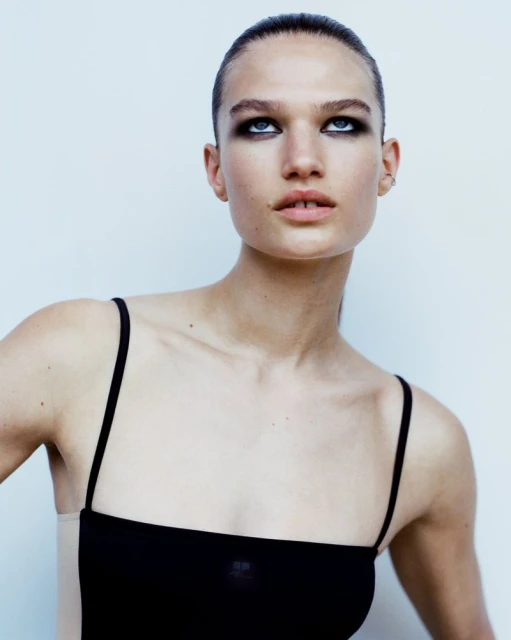 Portrait of a young adult female fashion model in a simple black top, fair skin, and dark eyeshadow against a plain white background.