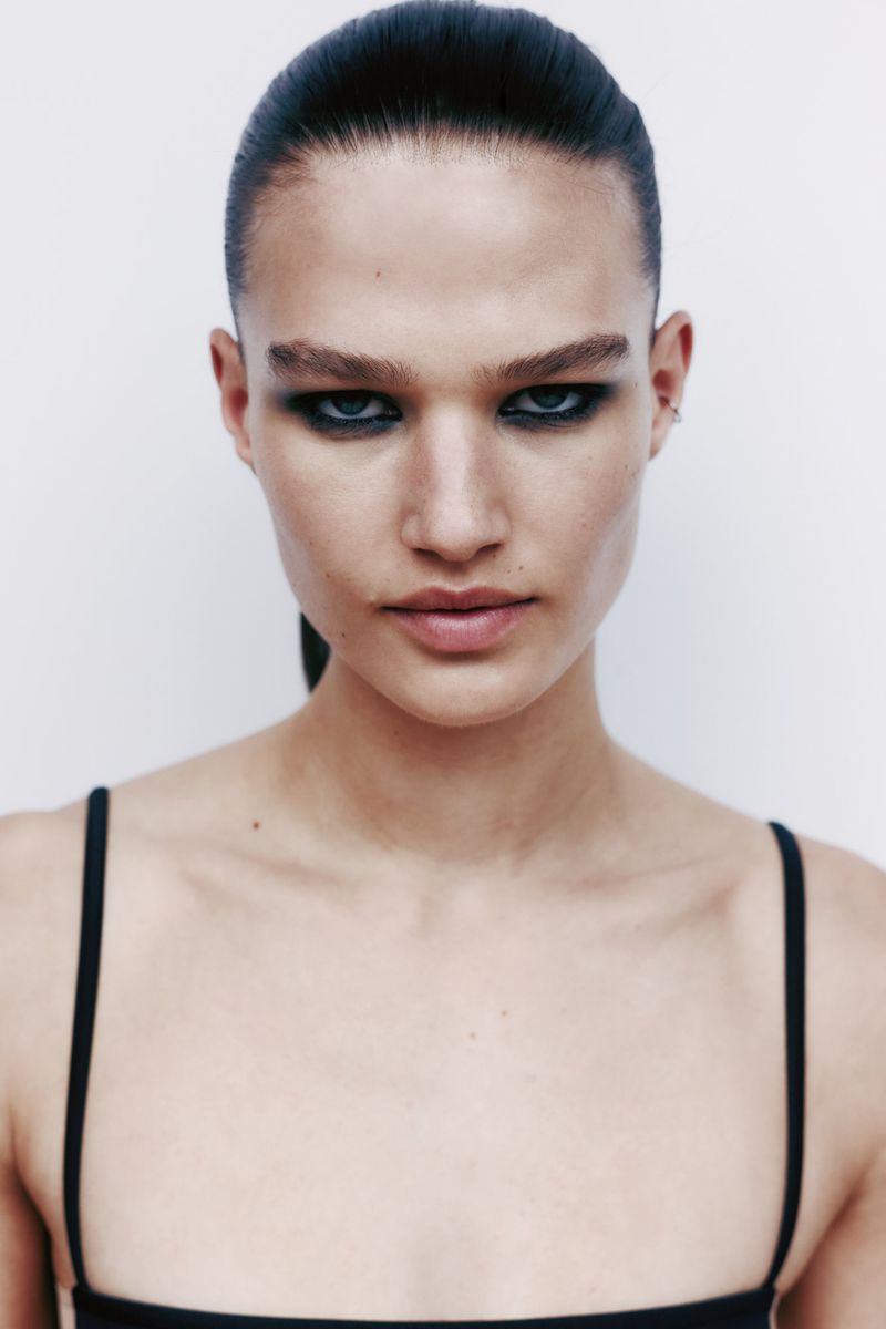 Close-up portrait of a person with dark eye makeup and slicked-back hair against a plain white background in a beauty or fashion context.