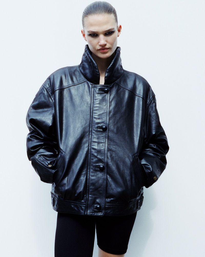 Portrait of an adult female fashion model posing in a black leather jacket and black bike shorts in a brightly lit photo studio.
