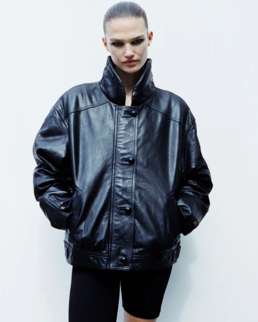 Portrait of an adult female fashion model posing in a black leather jacket and black bike shorts in a brightly lit photo studio.