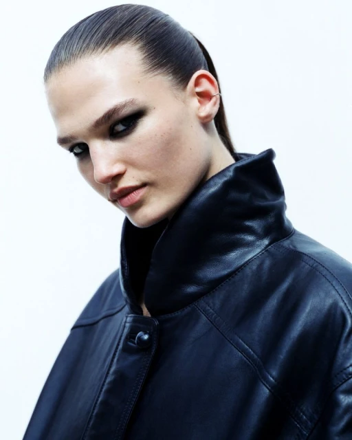 Close-up portrait of a woman with slicked-back hair and smokey eye makeup, wearing a dark, structured leather jacket against white backdrop.