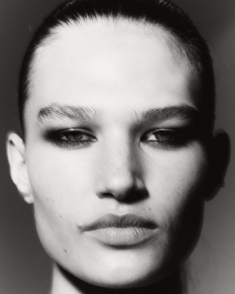 Close-up black and white portrait of a woman with sleek hair and strong makeup, focusing on her face and features in a neutral setting.