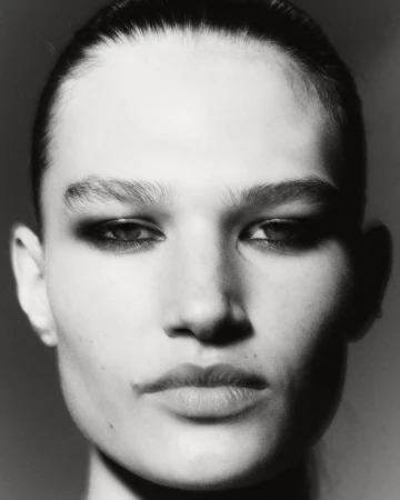 Close-up black and white portrait of a woman with sleek hair and strong makeup, focusing on her face and features in a neutral setting.