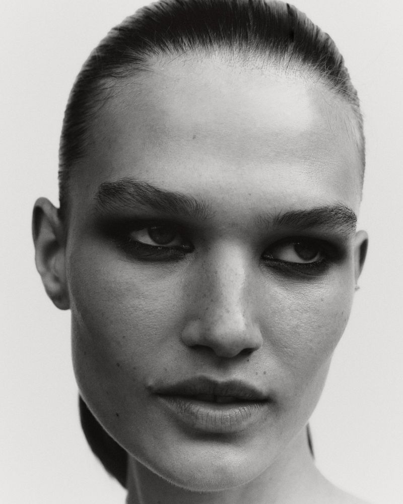 Monochrome portrait of a woman with slicked-back hair and dramatic eye makeup in a studio setting. High-contrast, beauty shot.