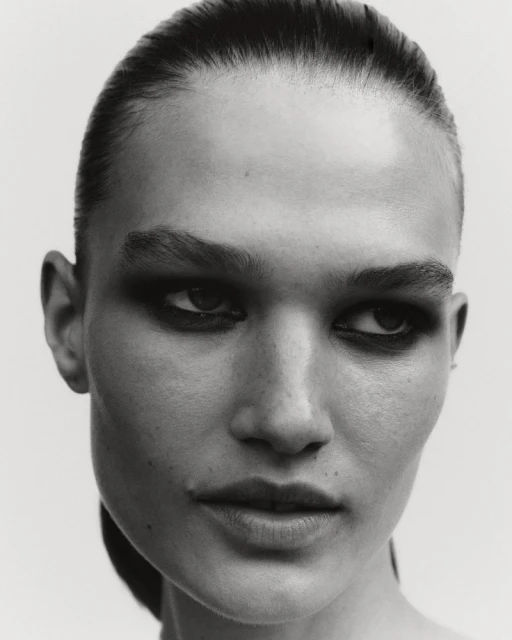 Monochrome portrait of a woman with slicked-back hair and dramatic eye makeup in a studio setting. High-contrast, beauty shot.