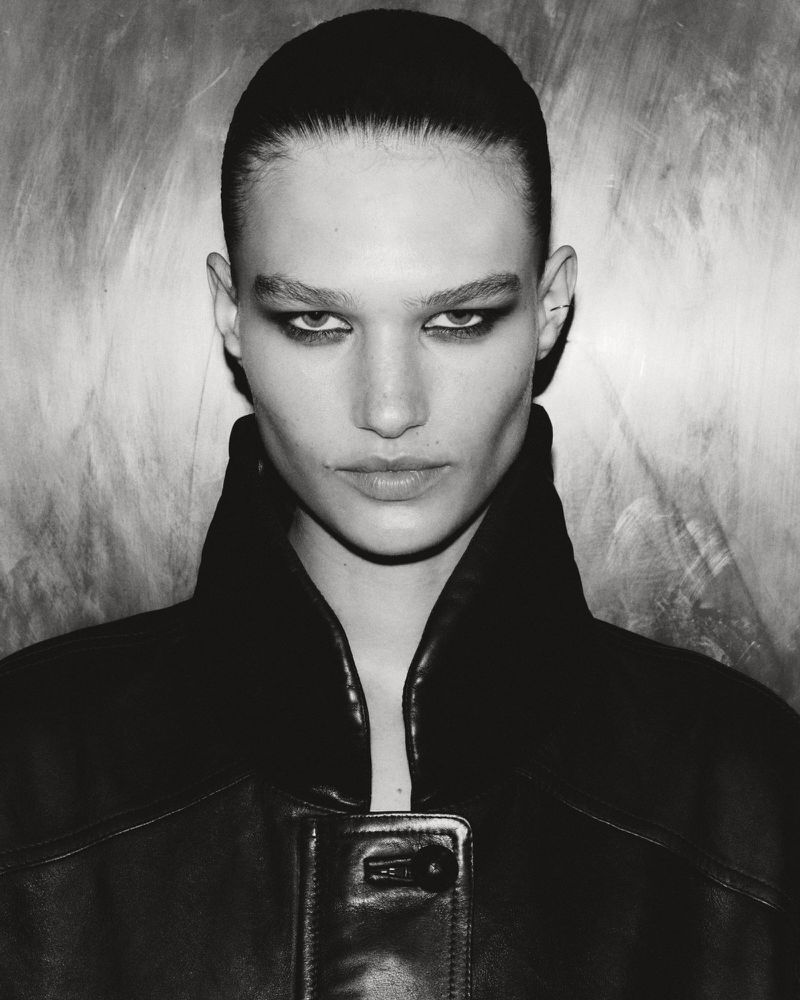 Monochrome portrait of a woman with sleek hair and dark makeup, wearing a black leather jacket against a textured background.