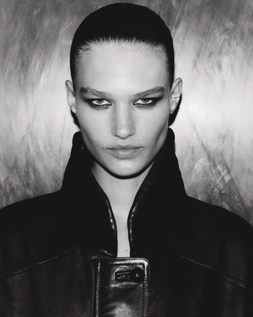 Monochrome portrait of a woman with sleek hair and dark makeup, wearing a black leather jacket against a textured background.