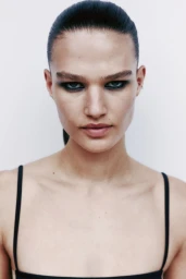 Emmi Freeman with slicked-back hair and dark smoky eye makeup wearing a black strap top against a clean white backdrop.
