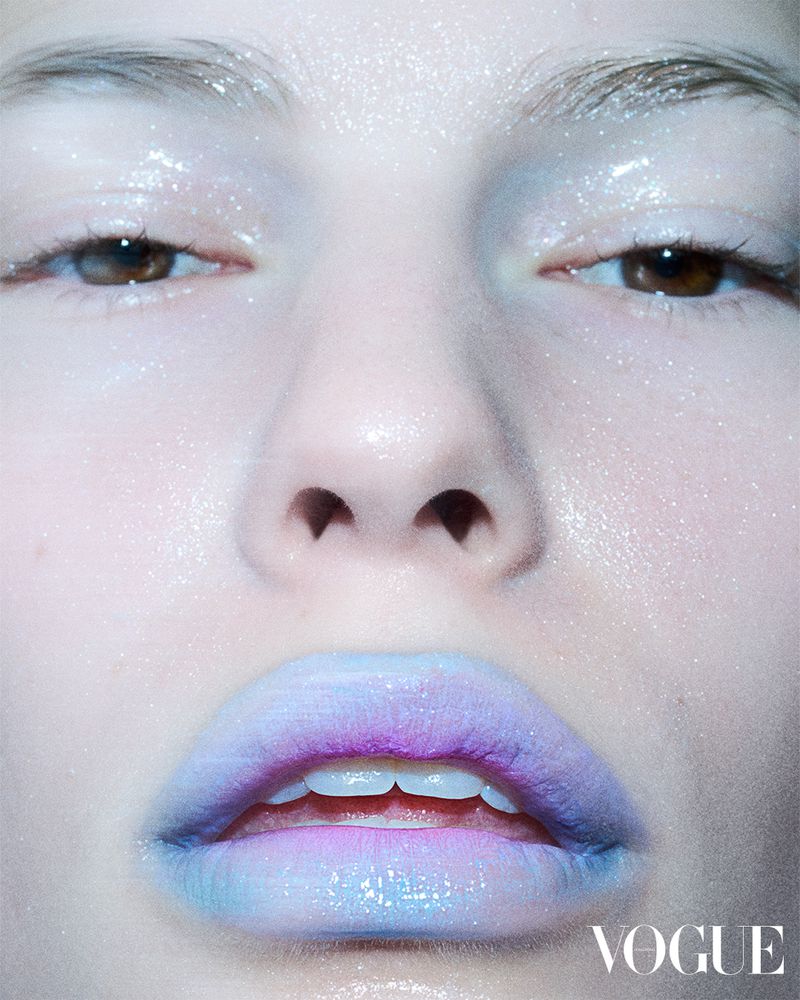 Close-up portrait of a woman's face with glossy, multi-colored lips. The model is lit with soft, diffused light, Vogue logo in corner.