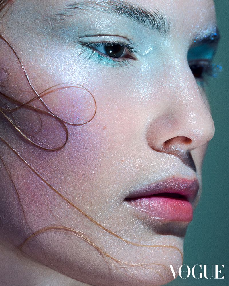 Close-up beauty shot of a fair-skinned person with bold makeup and wet strands of hair, set against a soft teal backdrop.