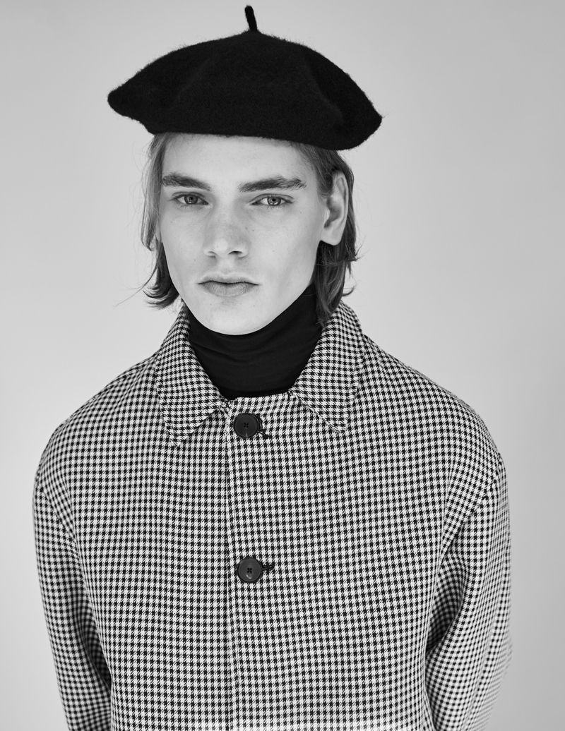 Black and white portrait of a young male model wearing a black beret, turtleneck, and patterned jacket looking at the camera.