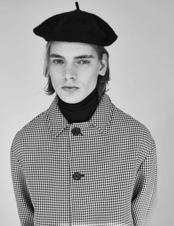 Black and white portrait of a young male model wearing a black beret, turtleneck, and patterned jacket looking at the camera.