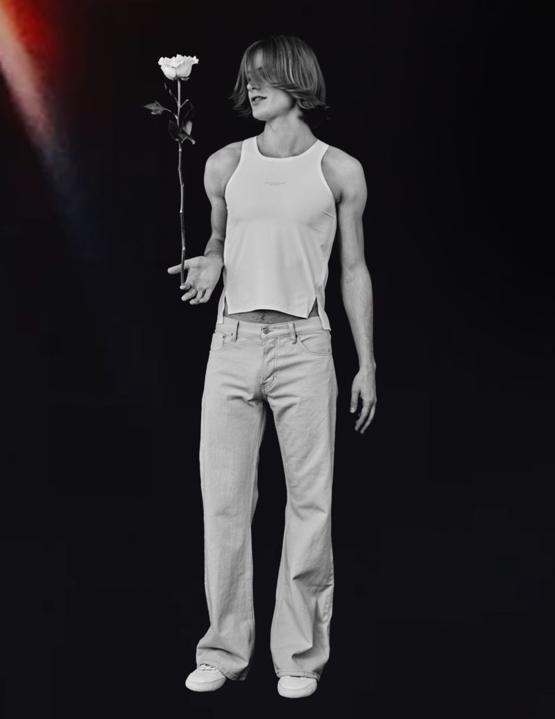 Studio shot of a young adult man holding a white rose, wearing a tank top and jeans, against a solid black background with red light.