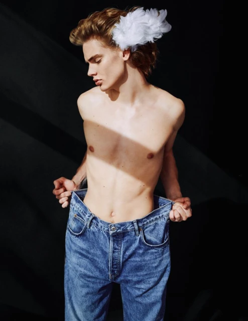 A young, blonde male model poses shirtless, holding large blue jeans against a dark background with a feathered hair accessory. 