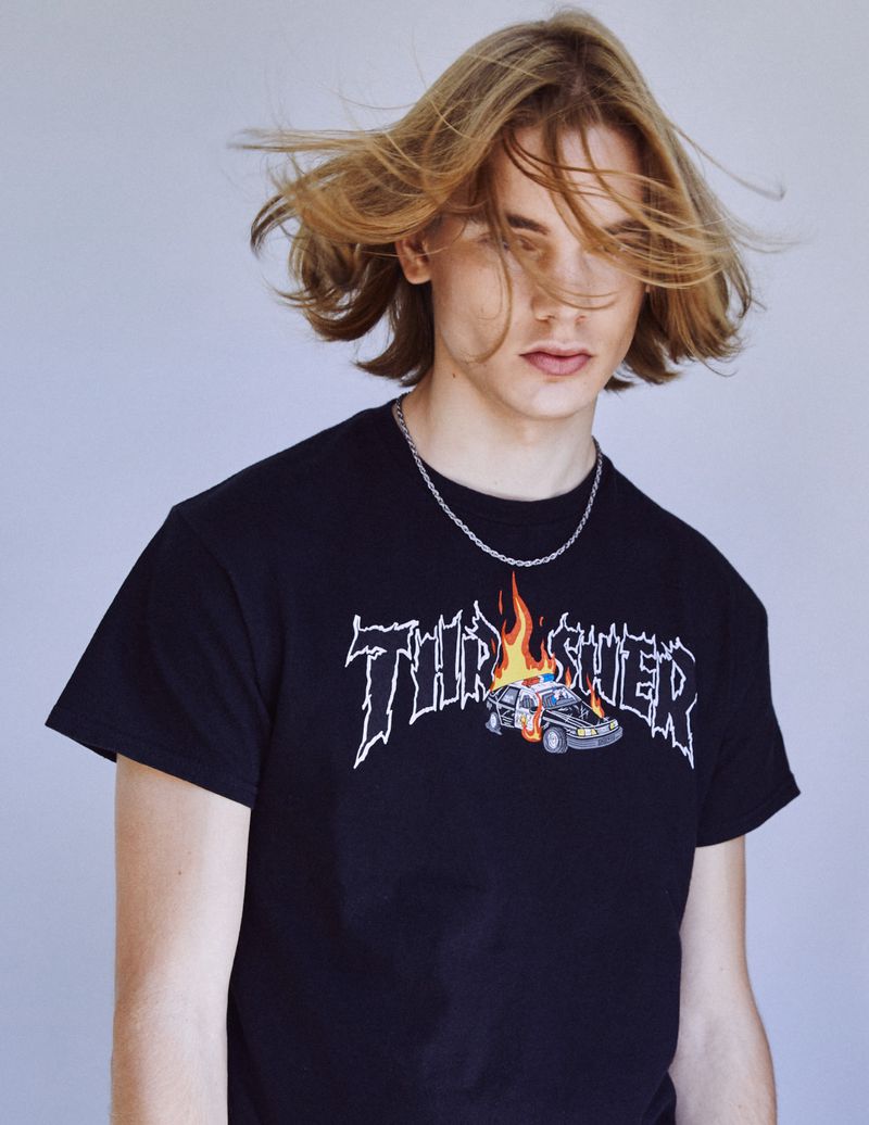 Young adult with flowing blonde hair wears a Thrasher logo T-shirt with a flaming police car graphic against a solid grey background.