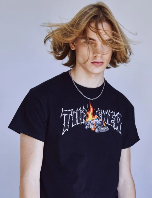 Young adult with flowing blonde hair wears a Thrasher logo T-shirt with a flaming police car graphic against a solid grey background.