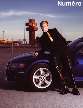 Model poses with a blue BMW near an airport control tower at sunset. Wearing a black suit and golden tie, fashion editorial shot.