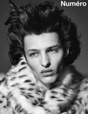 Black and white portrait of a male model with dark wavy hair wearing a fur coat in a studio setting with the text 'Numéro' visible.