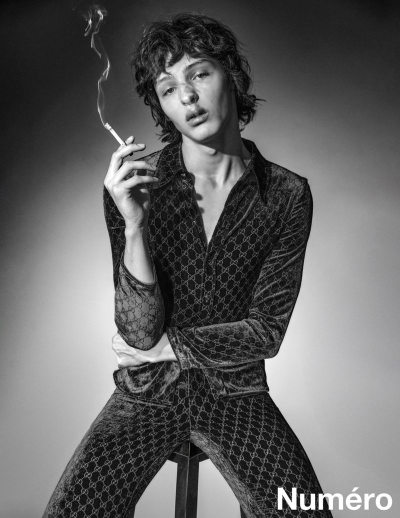 Monochrome portrait of a young adult male wearing a patterned velvet suit while smoking, captured in a studio setting.