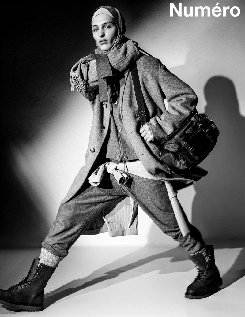 Fashion editorial shot of a person in a grey coat, scarf, and boots with a dark bag, in a studio with magazine logo text.