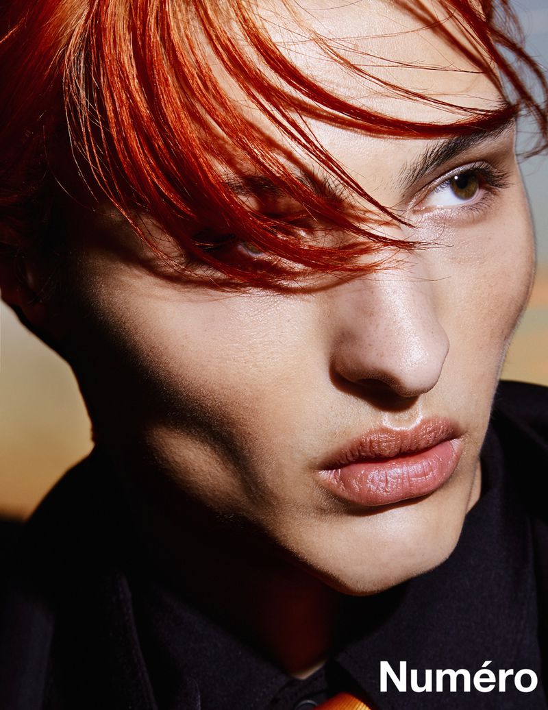 Close-up portrait of a person with red-orange hair and brown eyes, wearing a black shirt, against a blurred orange and blue background.
