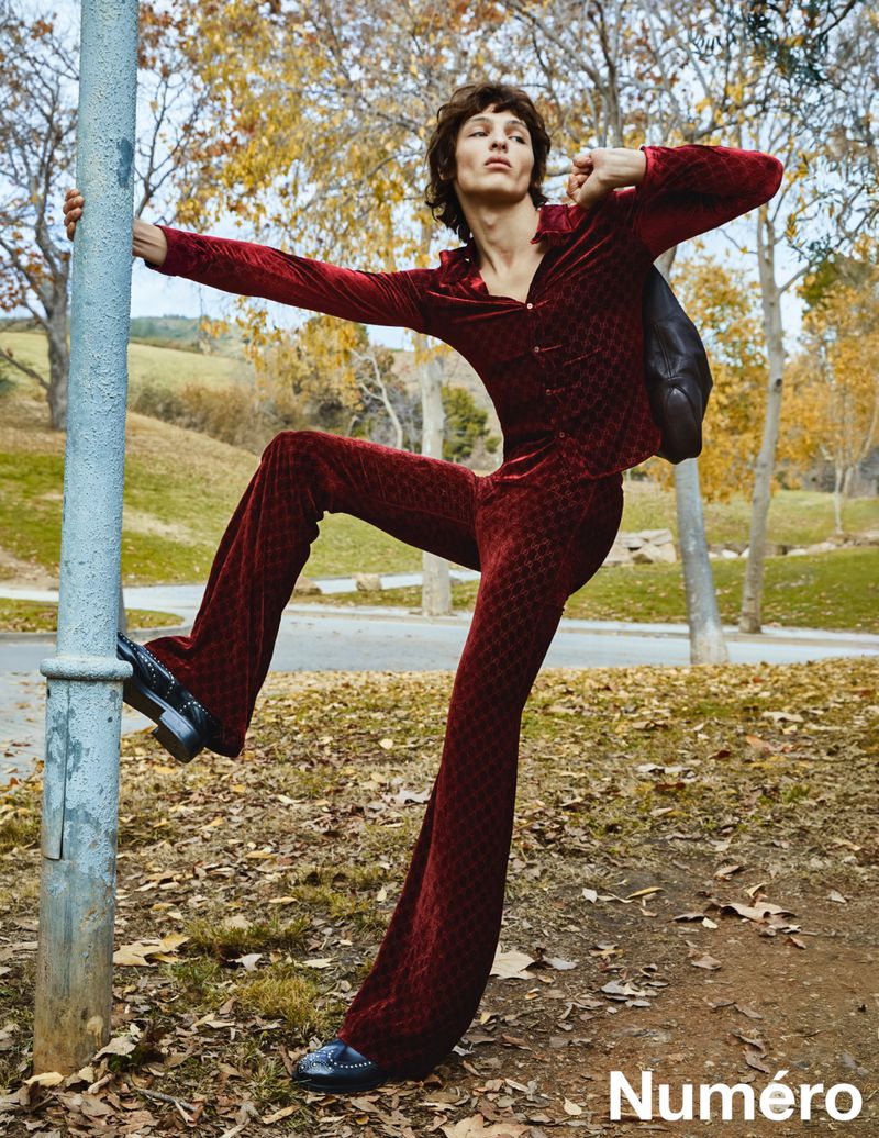 Man in velvet suit poses dynamically against a pole in an outdoor park setting, showcasing fashion-forward editorial style.