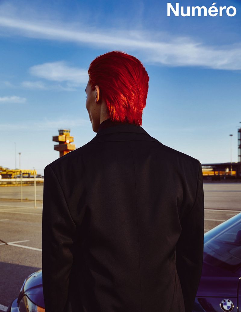Male model with red hair and black suit jacket stands with his back to the camera near a dark sedan and industrial buildings.