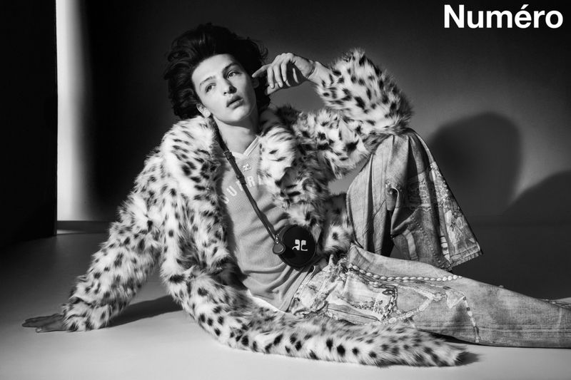 A young adult male fashion model reclines on the floor in a faux fur coat and denim jeans in a black and white editorial shot.