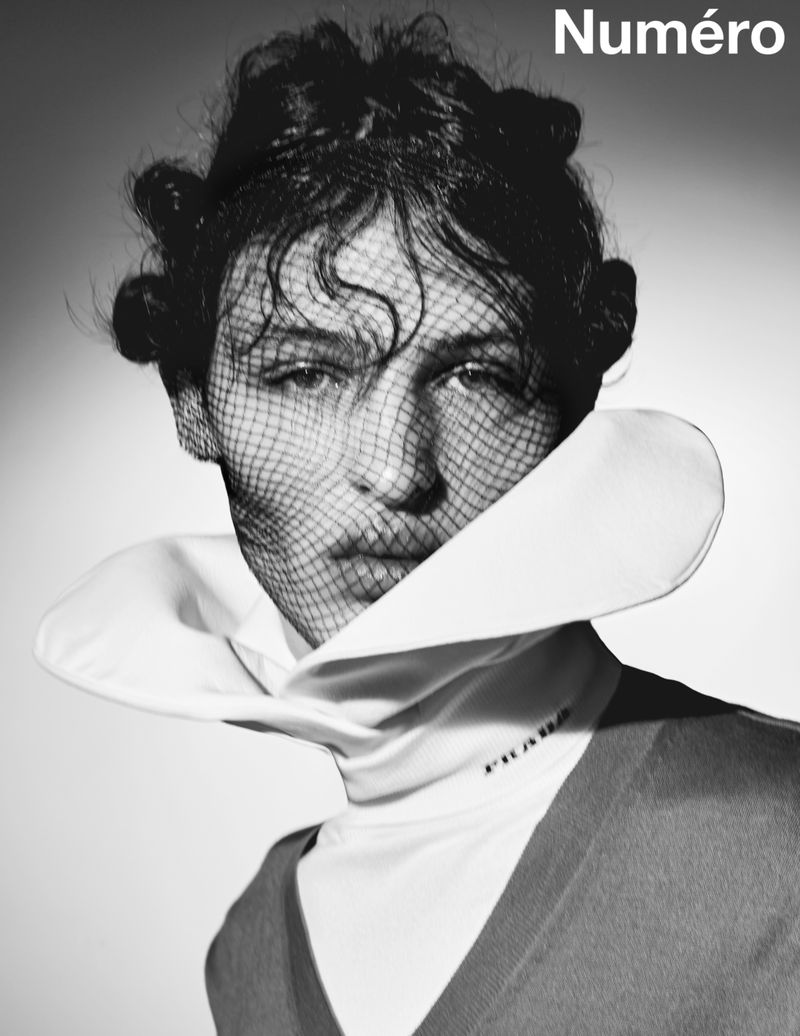 Monochrome portrait of a woman wearing a net veil and collared shirt with 'Prada' logo, set against a clean grey background.