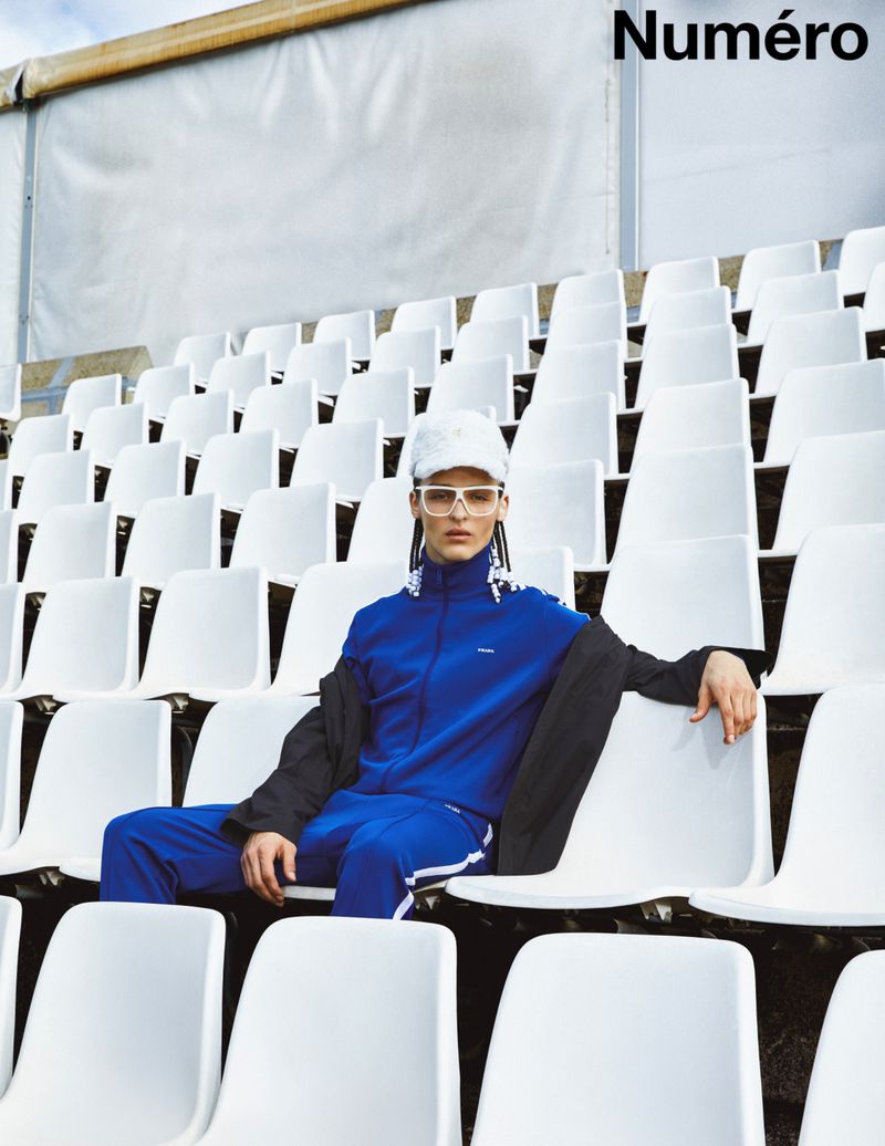 A person in a blue tracksuit sitting in stadium seating wearing sunglasses and a hat, with the magazine name 'Numéro' visible.