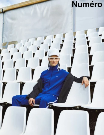 A person in a blue tracksuit sitting in stadium seating wearing sunglasses and a hat, with the magazine name 'Numéro' visible.