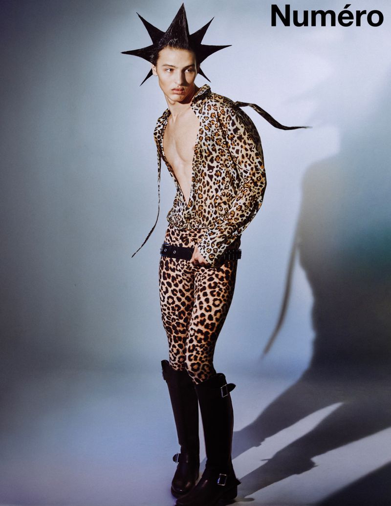 Fashion editorial featuring a male model in a cheetah-print outfit and spiky hairstyle, standing in a studio with soft, diffused lighting.
