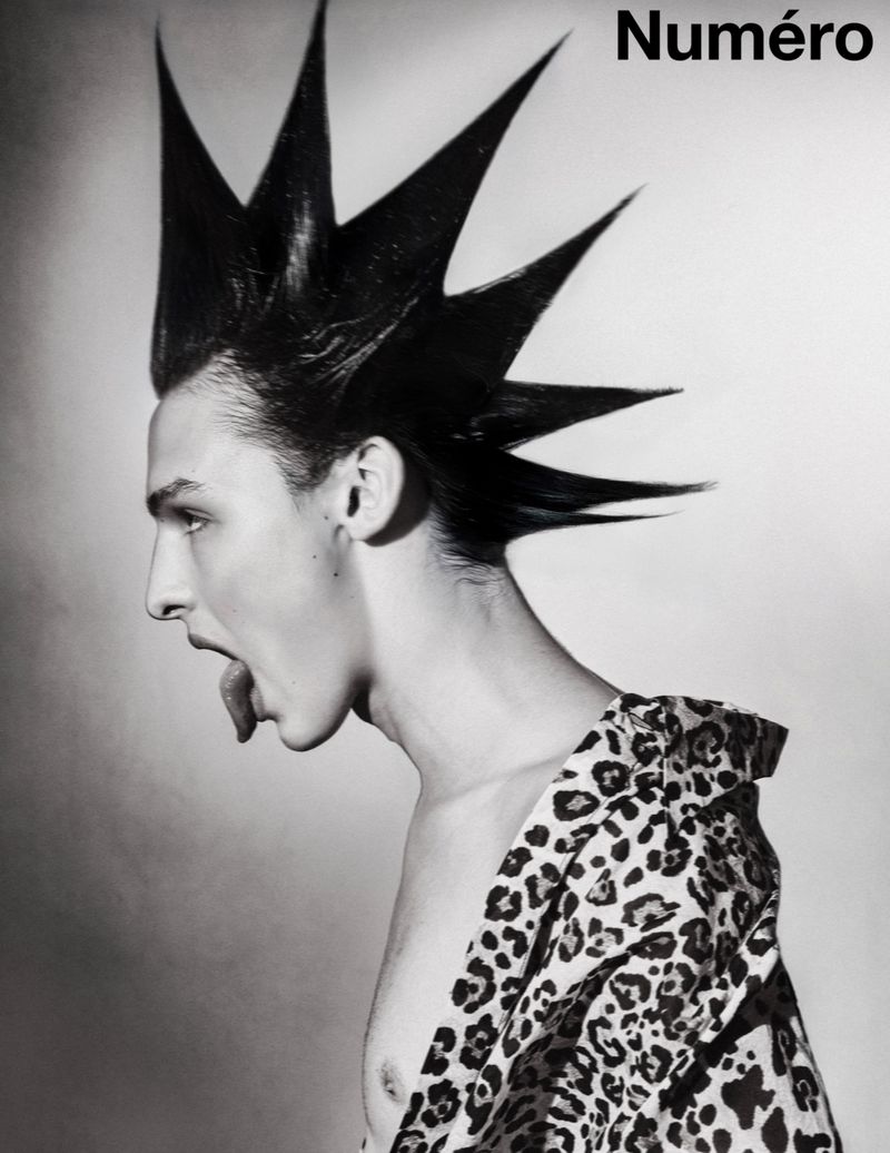 Monochrome portrait of a male model with a tall, spiky mohawk and an animal-print jacket, captured in a studio setting.