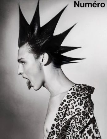 Profile of a man with dramatic spiked punk hair sticking his tongue out, wearing a leopard print shirt against a studio backdrop.