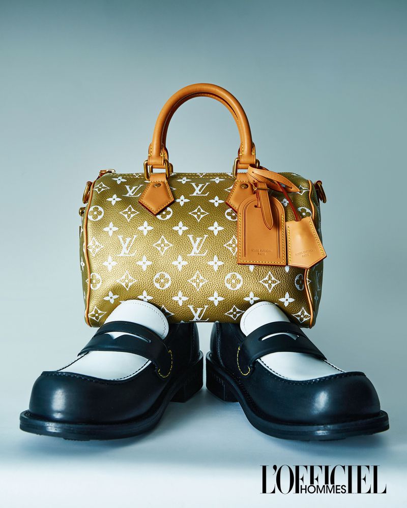 Louis Vuitton handbag in monogram canvas with leather trim, paired with black and white leather loafers against a solid grey backdrop.