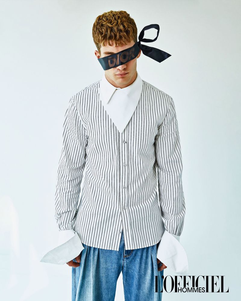 Male fashion model poses in a striped shirt and jeans, with a black ribbon blindfold. Studio lighting. L'Officiel Hommes logo visible.