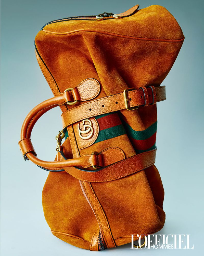 Tan Gucci duffel bag with leather straps and gold logo detail on a light blue background in a studio product shot for L'Officiel Hommes.
