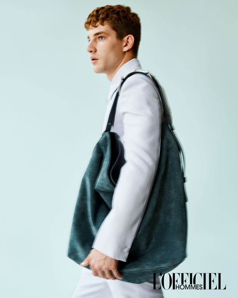 Fashion editorial shot of a young male model wearing white suit and holding a green tote bag against a light blue studio backdrop.