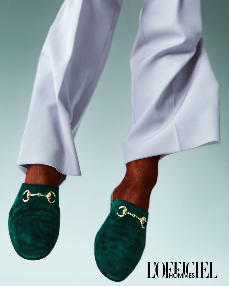 Elevated shot of a person wearing white trousers and green velvet Gucci Princetown mules, in a fashion studio setting with soft, diffused lighting.