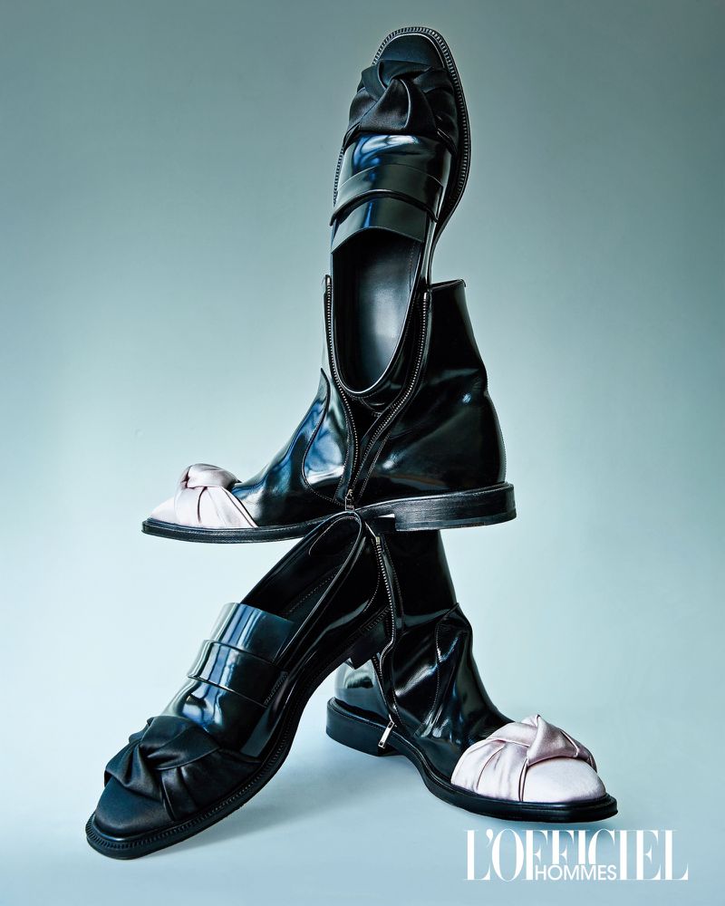 Fashion editorial shot of three black patent leather shoes stacked on each other against a light blue background, showcasing their design.