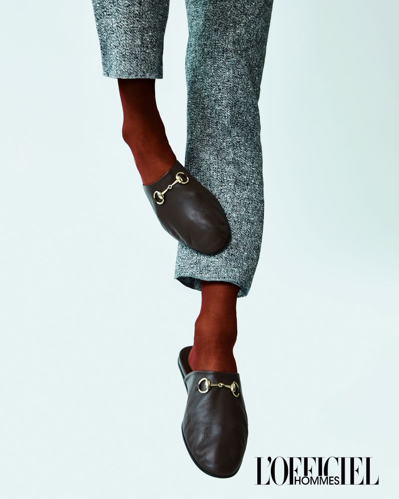 Eye-level shot of brown leather mules on a person wearing brown socks and grey tweed dress pants against a white backdrop.