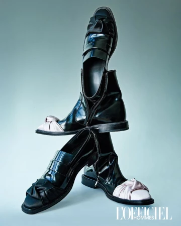 Sculptural stack of black patent leather boots, loafers, and satin knot shoes arranged vertically against a pale blue studio backdrop.