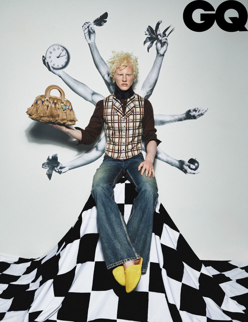 Fashion editorial shot: a light-skinned male model with blonde hair sits atop a black-and-white diamond pattern with a purse.