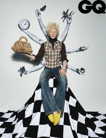 Fashion editorial shot: a light-skinned male model with blonde hair sits atop a black-and-white diamond pattern with a purse.