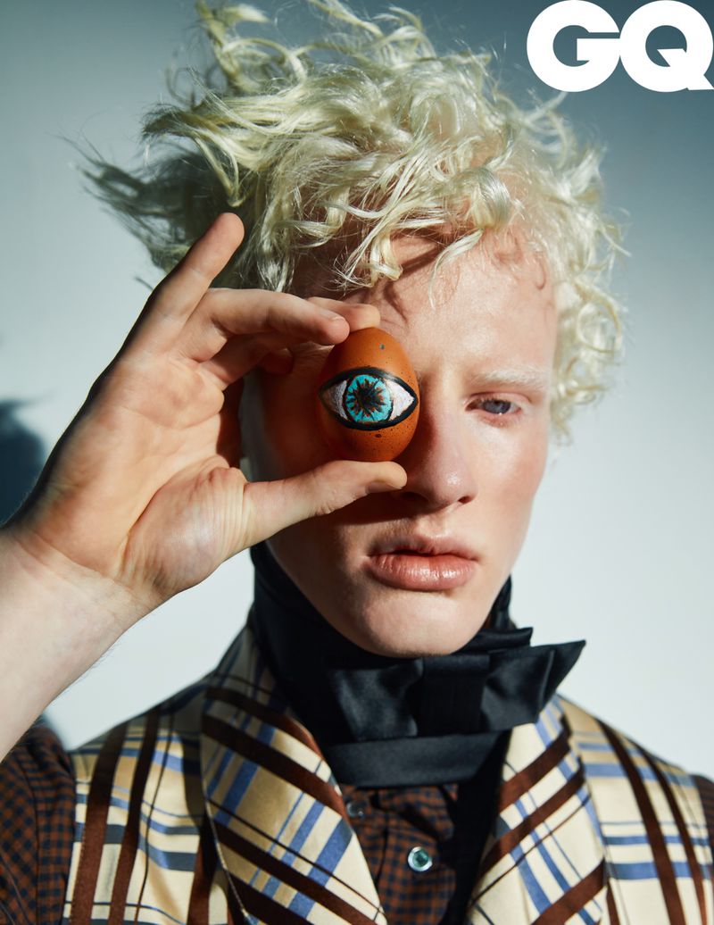 Fashion portrait of a fair-skinned man holding a painted egg over his eye, wearing a plaid vest and black ascot, in front of a white backdrop.