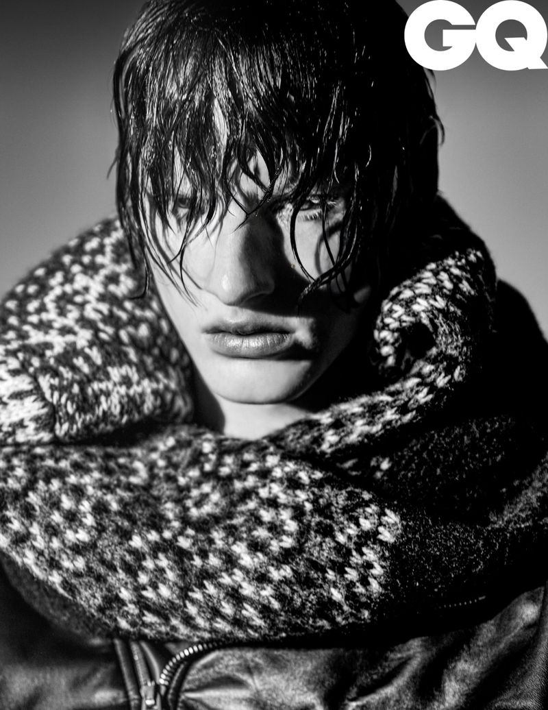 Monochrome portrait of a male model with wet hair wearing a chunky patterned knit scarf and leather jacket against a grey background.